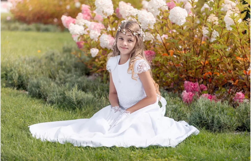 Urocza dziewczynka siedząca w ogrodzie w albie komunijnej sukienkowej typu sukienka, ozdobionej piękną bawełnianą koronką.