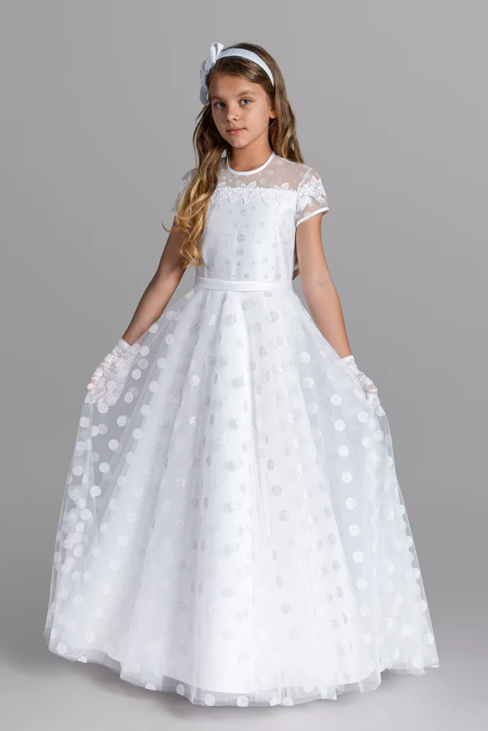 A girl in a communion dress with a polka dot tulle skirt. The dress has a delicate sleeve that adds to its charm. The dress is decorated with beautiful, delicate applications that beautifully emphasize its charm. The picture shows a girl in a charming communion dress. The skirt of the dress is made of tulle with a polka dot pattern, and its uniqueness is emphasized by a delicate sleeve. The dress is also decorated with beautiful applications that add to its unique charm. Discover the magic and elegance of this communion outfit for girls.
